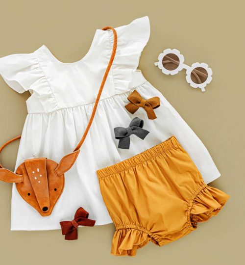 White dress,  orange shorts with kids handbag and sunglasses. Set of  baby clothes and accessories for spring or summer on beige background. Fashion childs outfit. Flat lay, top view